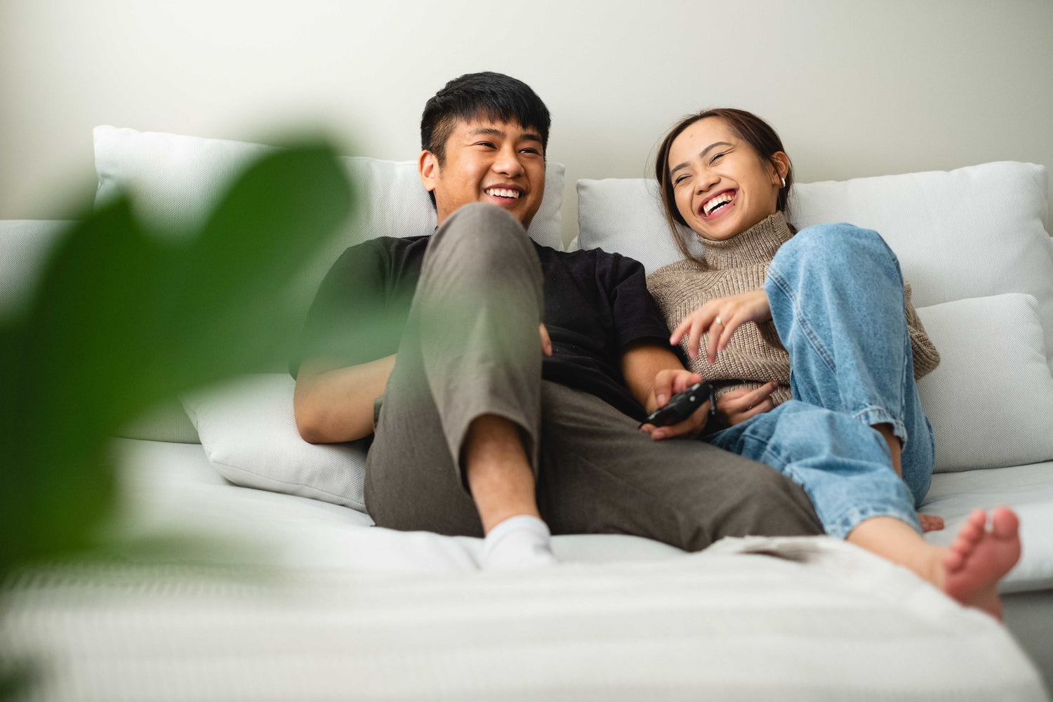Mann und Frau asiatischer Herkunft sitzen lachend auf einem Sofa und scheinen großen Spaß zu haben. Beide sind mit Kleidung angezogen außer der Frau. Die Frau trägt keine Socken.
